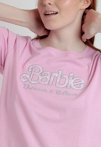 Camiseta de algodón rosa con mangas cortas que presenta el texto bordado en plata "Barbie Dreamer & Believer" en el pecho.