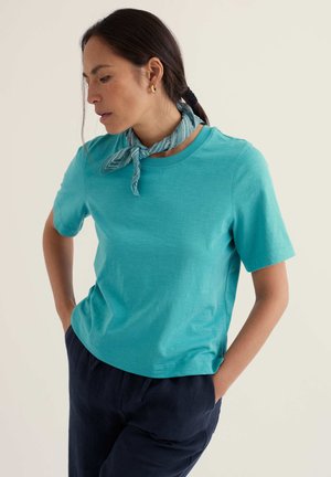 Sötét hajú, hátul összefogott hajjal rendelkező nő, arany karika fülbevalóval, kékeszöld rövid ujjú ingben, tengerészkék nadrágban és világoskék mintás nyakkendőben, lefelé nézve.