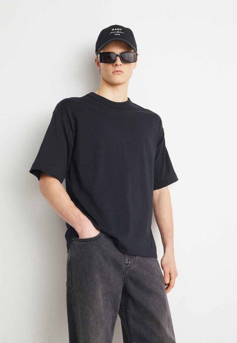 T-shirt nera in cotone con maniche corte e scollatura tonda; abbinata a pantaloni in denim grigi e un cappellino nero con logo bianco e occhiali da sole.