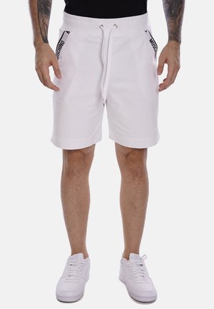 Pantaloni corti bianchi in cotone con cordino, due tasche laterali, accenti neri del logo sui lati e una vestibilità rilassata. Indossati con scarpe da tennis bianche.