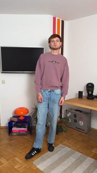 Világos lila pulóver grafikus részletekkel, világoskék farmerral párosítva. Fekete sportcipők és tetoválások láthatók. Belső tér dekorációval.