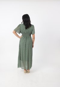 Maxikleid aus grünem Stoff mit floralem Muster, gerafftem Taillenbereich, kurzen Ärmeln und fließender Silhouette. Stil kombiniert mit hellen Schuhen.