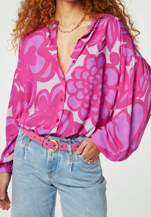 Femme portant un chemisier floral rose et violet avec des manches bouffantes, un jean bleu clair et une ceinture rose avec une boucle dorée.