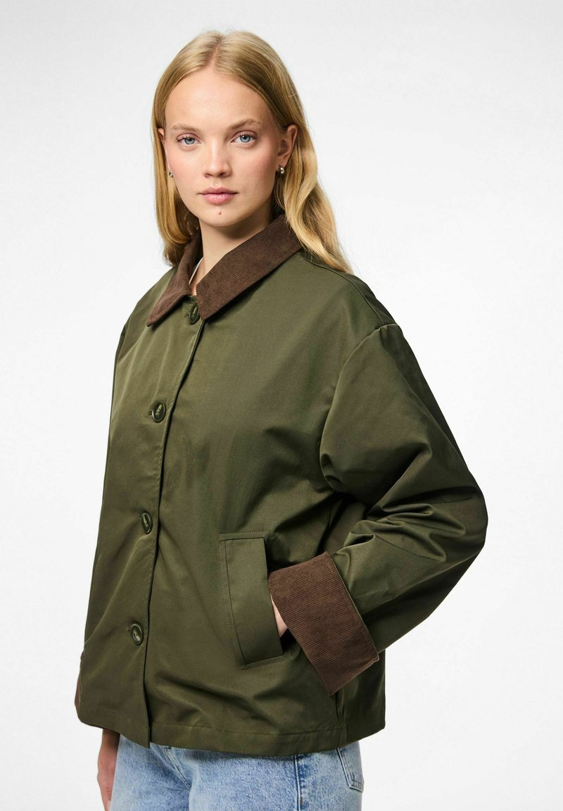 Jauna moteris vilki alyvuogių žalios spalvos striukę su rudomis apykaklėmis ir rankogaliais, šviesias džinsines kelnes, pozicionuojasi su viena ranka kišenėje prieš baltą foną.