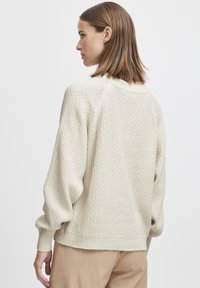 Pull en tricot crème avec un motif texturé, des manches raglan et des poignets côtelés. La vue de dos montre une coupe décontractée et un col rond.