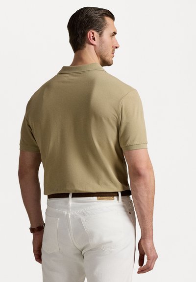 Κεραμιδί κοντομάνικο πόλο μπλουζάκι με υφή και γιακά, συνδυασμένο με λευκά παντελόνια και καφέ ζώνη, που φαίνεται από πίσω.