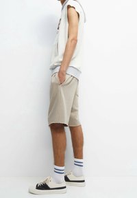 Light gray sleeveless hooded sweatshirt with a pocket, paired with beige shorts featuring a frayed hem. White socks with blue stripes and sneakers.