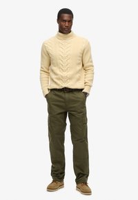 Romige kabelgebreide coltrui, olijfgroene cargo broek en beige veterschoenen. Model staat met handen in de zakken tegen een effen achtergrond.