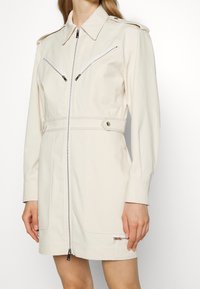 Světle béžové užitkové šaty se zipem vpředu, sklopeným límcem, dvěma kapsami na hrudi a patentkami na ramenech; hladká tkanina s přizpůsobeným střihem.