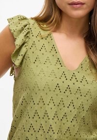 Grøn bomuldsbluse med en dyb V-hals, flæser på ærmerne og et struktureret mønster af udskæringer, der danner et zigzag-design.