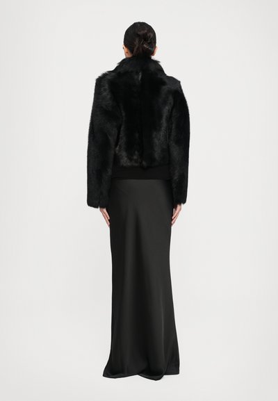Woman with dark hair in a bun wearing a black faux fur jacket and a long black skirt, standing against a plain light background.