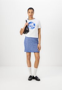 Hvid grafisk t-shirt med blåt design, parret med en blå mønstret miniskørt. Modellen bærer sorte loafers og knæhøje hvide strømper.