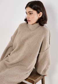 Beige übergroßer Pullover aus weichem Strickmaterial, mit hohem Kragen und überschnittenen Schultern. Glatte Textur mit geripptem Kragen-Detail.