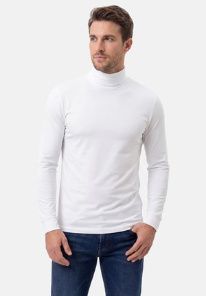 Mann mit kurzen braunen Haaren, der einen weißen Langarmrollkragen und blaue Jeans trägt und vor einem schlichten, hellen Hintergrund steht.