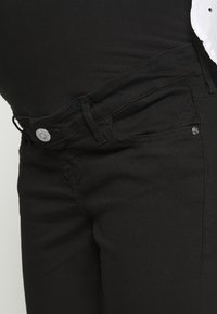 Jeans de denim preto com fecho padrão de botão, dois bolsos frontais e um design ajustado com detalhes de costura visíveis.