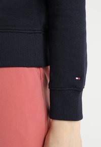Sudadera azul marino con puños acanalados, costuras visibles y un pequeño parche de logo en la dobladillo que presenta colores rojo, blanco y azul. Combinada con tela rosa.
