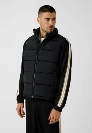 Homme portant une veste matelassée noire sur un sweat-shirt noir avec des rayures beiges et blanches aux manches, regardant sur le côté sur un fond uni.