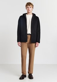 Giovane uomo in piedi che indossa una giacca nera con zip, un maglione color crema, pantaloni marroni, una cintura nera e scarpe marrone scuro su uno sfondo neutro.