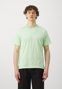 Lys grøn bomulds t-shirt med korte ærmer, rund hals og et hvidt logo centreret på brystet. Iført sorte bukser.