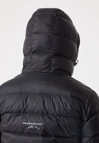 Svart uppblåsbar jacka med huva, featuring en quiltad design, slät yta, och logotyp på baksidan nära nacken. Justerbar dragsko.