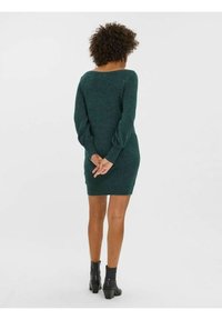 Grön stickad klänning med långa ärmar, smal passform och vid halsringning. Bärs med svarta ankelstövlar. Händerna ihopklämda bakom ryggen.