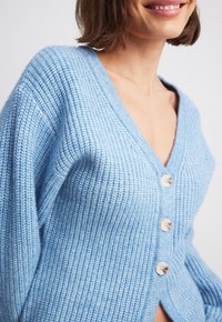 Světle modrý pletený kardigan s výstřihem do V a třemi předními knoflíky. Má texturovaný vzor a spadená ramena, které poskytují volný střih.