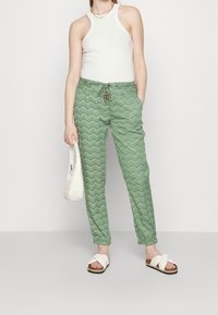 Woman wearing white sleeveless top, green zigzag patterned pants with drawstring, white sandals, and holding a small white handbag.