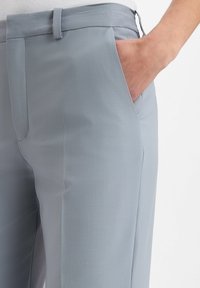 Nahaufnahme von hellblauen, maßgeschneiderten Hosen mit einer Hand, die in die vordere Tasche gesteckt ist, vor einem weißen Hintergrund.