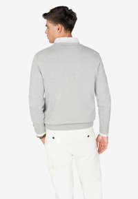 Jersey gris de punto con puños y dobladillo acanalados, llevado sobre una camisa blanca de cuello, combinado con pantalones de color claro. Vista trasera.