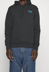 Svart bomullshuvtröja med en framficka och dragsko på huvan; har en liten, färgglad Nike-logotyp broderad på bröstet.