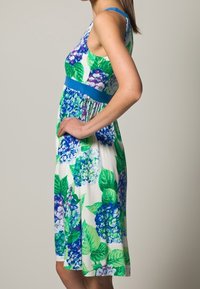 Kwiatowa sukienka bez rękawów z wzorem niebieskich i zielonych hortensji na białym tle, z jednolitą niebieską talią i długości do kolan.