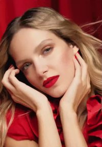 Nærbilde av et ansikt med blondt hår, røde lepper, og hender som forsiktig berører kinnene, mot en teksturert rød bakgrunn.