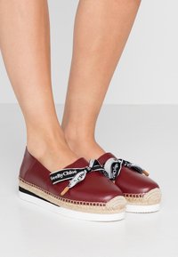 Espadrilles en cuir rouge avec une semelle en jute tissée, présentant des nœuds en ruban de marque noir et blanc à l'avant et une semelle intermédiaire blanche unie.