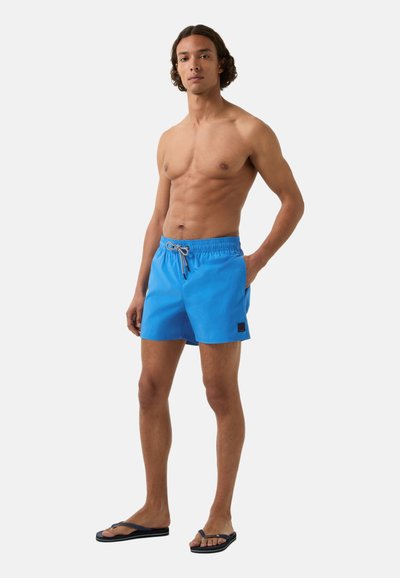 Modré plavky na plavání s elastickým pasem a šňůrkou. Obsahuje malou černou nášivku s logem. Model má na sobě černé žabky.