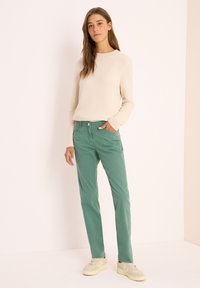 Suéter de punto texturizado color crema, combinado con pantalones rectos verdes y zapatillas claras. La modelo posa con una mano en el bolsillo.