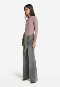 Kvinna med långt mörkt hår som bär en mauve långärmad topp, vida grå jeans, bruna skor och en grön tröja knuten runt midjan.