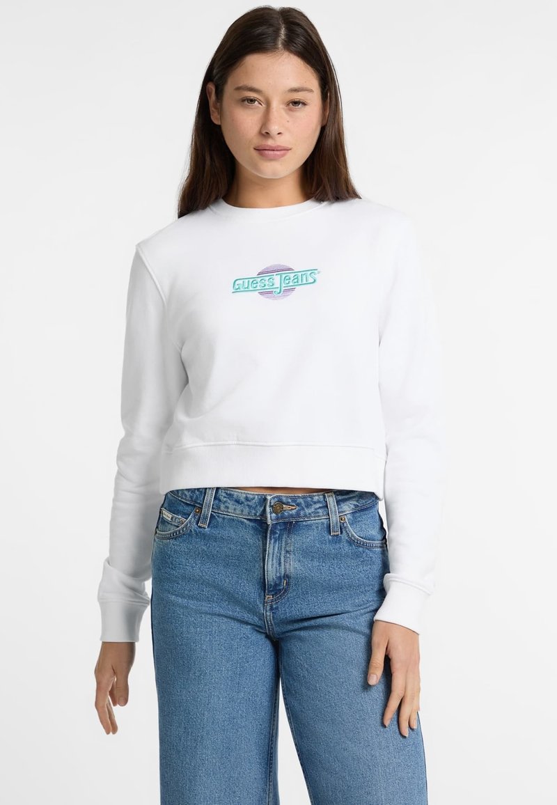 Valkoinen lyhyt huppari, jossa on ribbauskankaiset hihansuut, ja jossa on monivärinen "Guess Jeans" -logo keskellä rintaa, yhdistettynä sinisiin korkeavyötäröisiin farkkuihin.