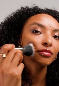 Femme aux cheveux bouclés appliquant du fond de teint avec un pinceau sur sa joue et son front, en se concentrant sur un teint uniforme et le maquillage.