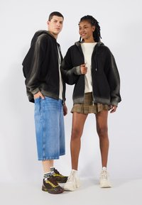 Felpe nere oversize con dettagli sfumati e accenti grigi chiari, abbinate a pantaloni corti in denim blu baggy e una gonna a quadri. Sneakers bianche e fantasia.