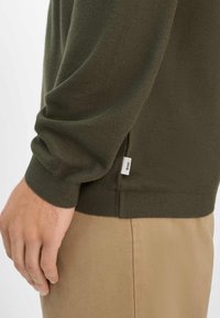 Olivgrüner, strukturierter Pullover mit gerippten Bündchen und Saum, versehen mit einem kleinen weißen Etikett an der Seite. Getragen über khakifarbenen Hosen.