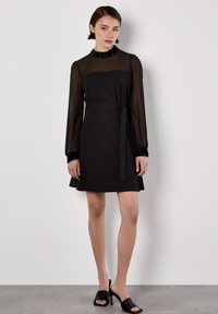 Vestido negro con cuello alto, mangas largas transparentes y cinturón de lazo. Acentos de cuentas en los puños. Textura suave, diseño hasta la rodilla.