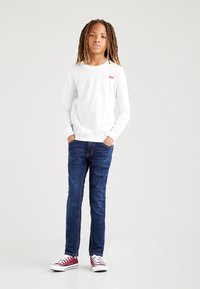 Bijela majica dugih rukava s crvenim Levi's logom, uparen s plavim trapericama i crvenim niskim tenisicama. Samostalni model s rukama u džepovima.