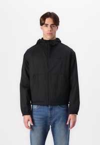 Svart hettejakke med glidelås i front, laget av lett, teksturert stoff, med elastiske mansjetter og to lommer foran. Brukt over blå jeans.