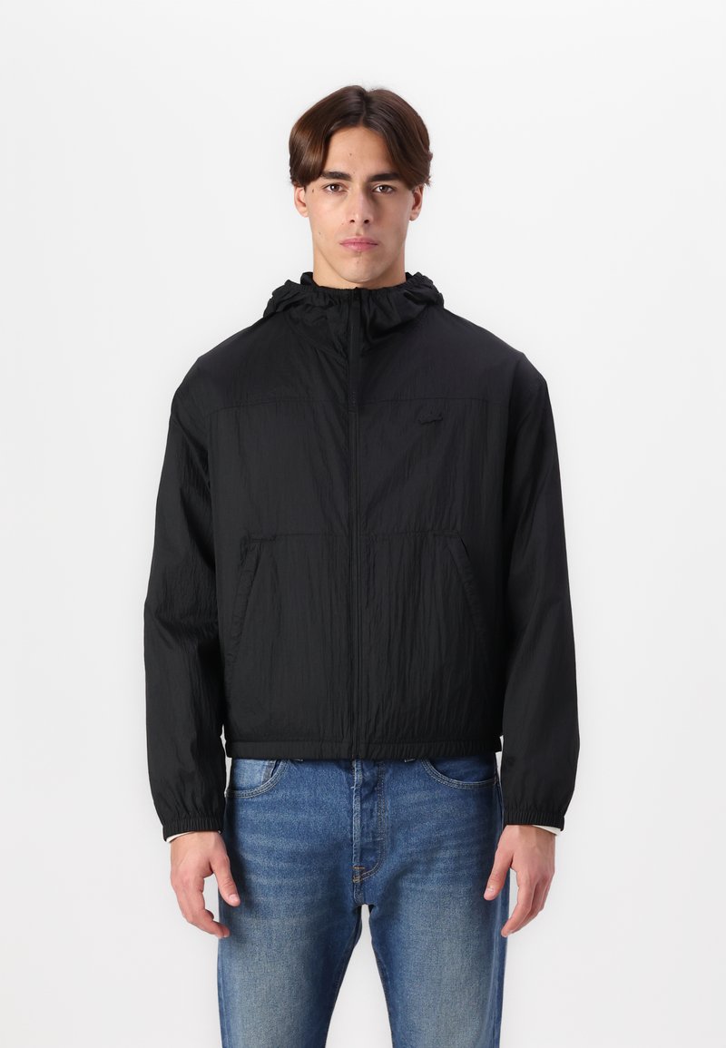 Svart hettejakke med glidelås i front, laget av lett, teksturert stoff, med elastiske mansjetter og to lommer foran. Brukt over blå jeans.