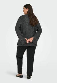 Donna con lunghi capelli castani, che indossa un maglione oversize grigio scuro con polsini borchiati, pantaloni neri e scarpe nere senza tacco, in piedi con le mani intrecciate dietro la schiena.
