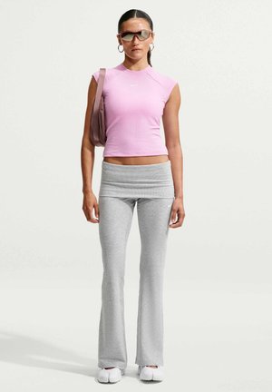 Femme portant un haut sans manches rose, des leggings évasés gris, des chaussures blanches, des lunettes de soleil et portant un sac à épaule mauve, debout devant un fond uni.