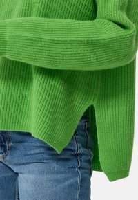 Grüner Strickpullover mit geripptem Design, lässiger Schnitt und seitlichen Schlitzen. Kombiniert mit blauen Denim-Jeans.