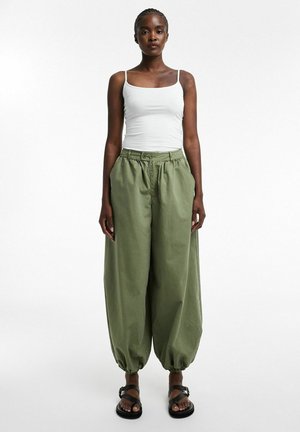 Vrouw draagt een witte tanktop, losse olijfgroene broek met aansnoerende enkels en zwarte sandalen, staand tegen een effen witte achtergrond.