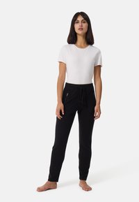 Schwarze Jogginghosen mit einem Kordelzugbund, ausgestattet mit zwei seitlichen Reißverschlusstaschen. Kombiniert mit einem schlichten weißen T-Shirt und barfuß.