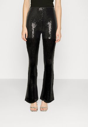 Femme portant un pantalon à paillettes noir taille haute avec jambes évasées et des sandales ouvertes noires et blanches à motifs, debout sur un fond blanc.